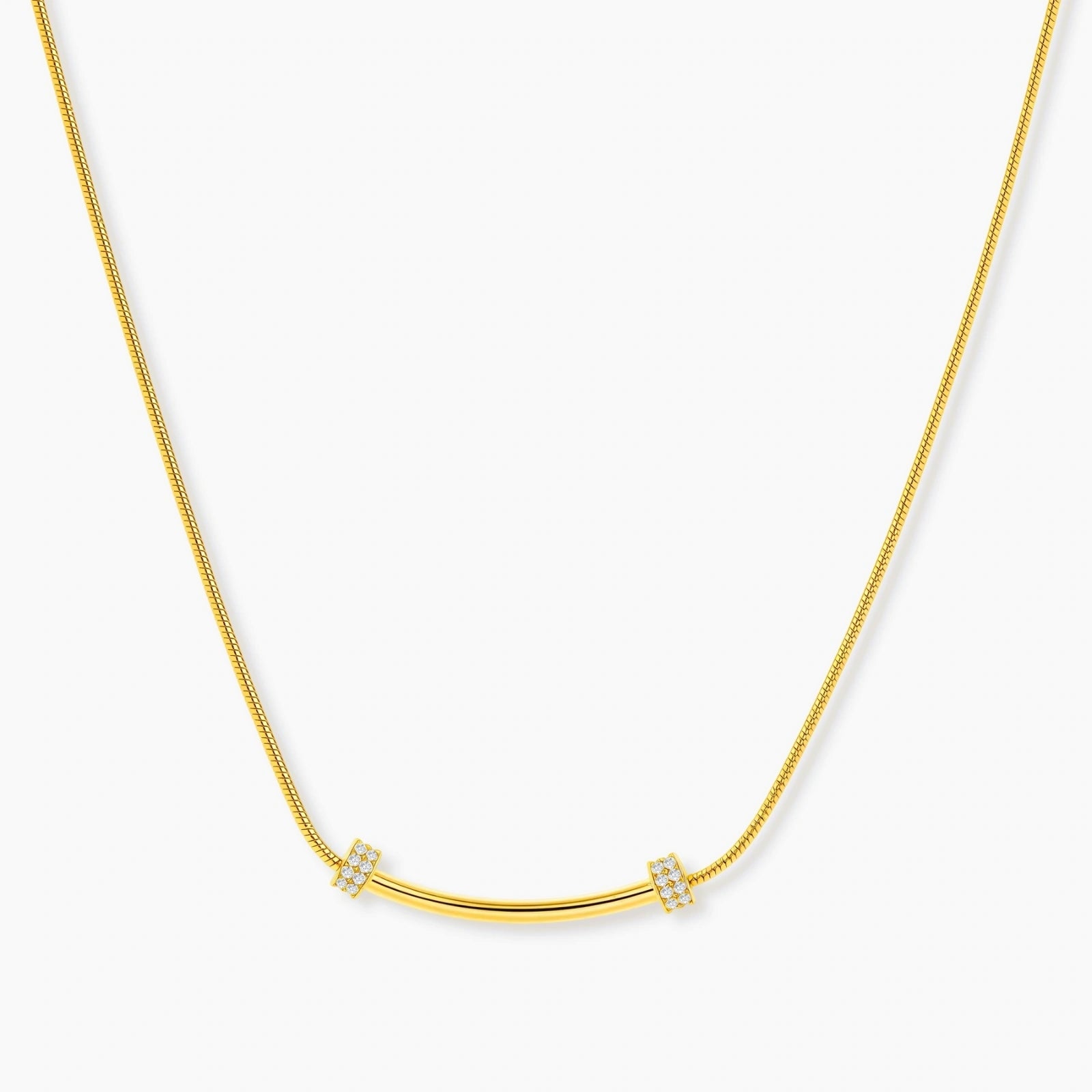 Delicate gold-plated necklace with curved bar pendant and crystal accents on snake chain against cream background.