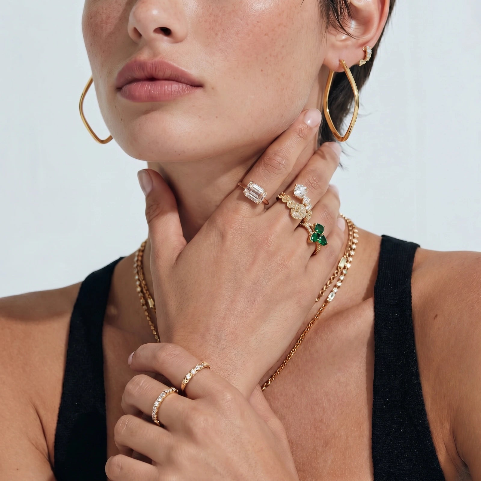 Close-up of a woman's face wearing gold hoop earrings, rings, and a necklace against a white background.